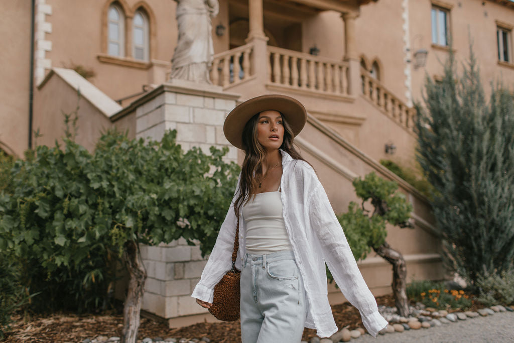 Roux Collective Boutique. Model wearing brown brim hat, white tank top with white button up over top, and light wash blue jeans.